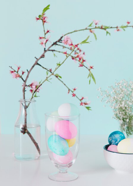 Яркие пасхальные яйца возле цветочной веточки в вазе с водой и миской