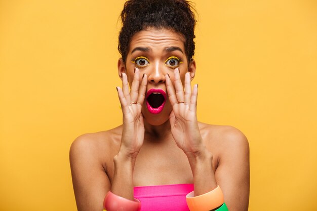 Яркая возбужденная стильная афроамериканская женщина, эмоционально кричащая или кричащая на камеру, кладущую руки в рот, изолированную над желтой стеной