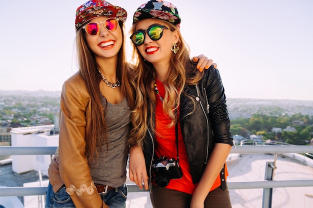 Free photo bright fashion outdoor lifestyle portrait of two pretty sisters wearing stylish swag hats, leather jacket and sunglasses, screaming laughing and having fun together.best fiends posing in the roof