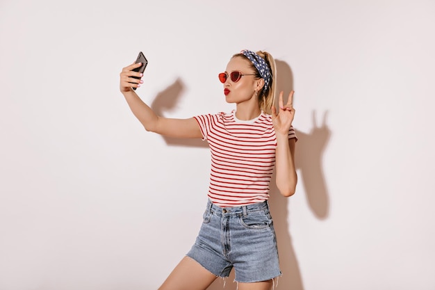 Bright girl with red lips and sunglasses in striped tshirt and denim cool shorts showing peace sign and making selfie on white background