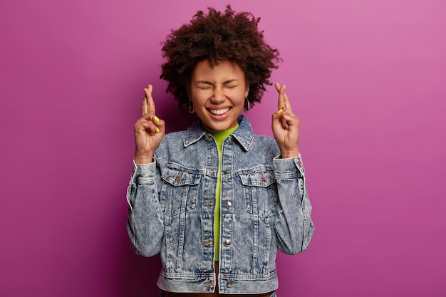 Bright image of happy overemotive woman with curly hairstyle, crosses fingers for good luck, believes in something pleasant happen, prays for better, keeps eyes closed, isolated over purple wall