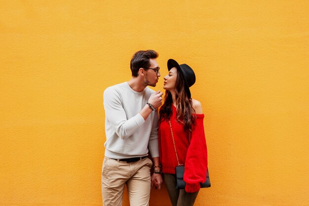 Bright image of lovers posing on yellow wall. Fashionable look. Romantic mood. Holding hands. Young woman with candid smile flirting with her boyfriend. Luxury bag.