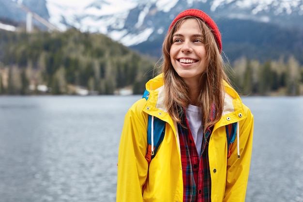 Bright image of young female traveler stands against mountain lake space, wears stylish red hat and yellow coat