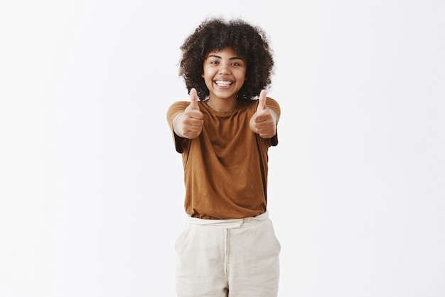 スタイリッシュな茶色のtシャツでアフロの髪型で明るく楽しい喜びのアフリカ系アメリカ人の女の子は親指で手を引っ張って、応援と支持を笑顔