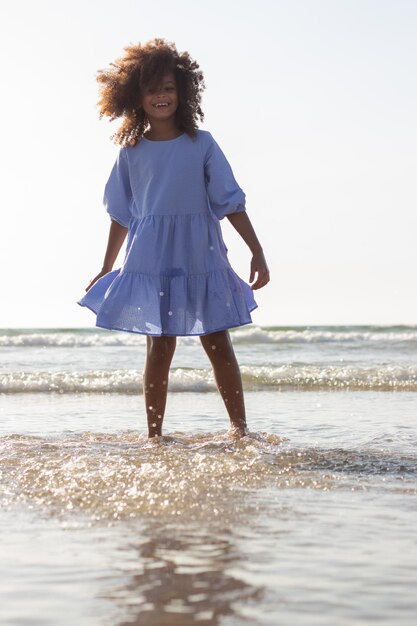Яркая маленькая девочка в платье на пляже. Афроамериканский ребенок на пляже в летний день, брызгая водой, улыбаясь. Детство, отдых, концепция счастья