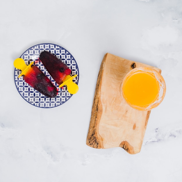 Free photo bright popsicles on plate and glass with juice on wooden stand on gray surface