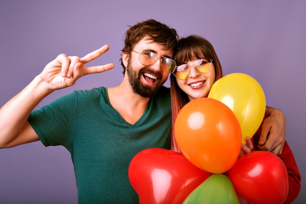 Bright  positive portrait of happy pretty couple smiling, showing peace gesture and holding party balloons, family relation, violet wall