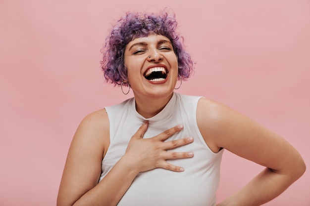 Bright positive woman with purple short wavy hair in white outfit and earrings with pierced nose laughs on pink.