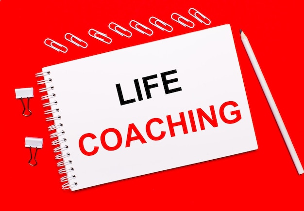 On a bright red background, a white pencil, white paper clips, and a white notebook with the text life coaching