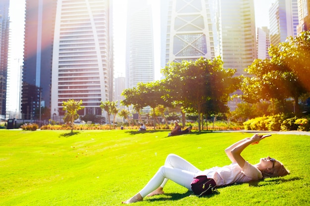 Bright summer sun shines over lady lying on green lawn and checking her Smartphone in the park