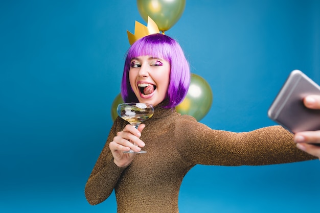 Celebrazione brillante emozioni di giovane donna con taglio di capelli viola che fa il ritratto di selfie. palloncini d'oro, divertirsi, mostrare la lingua, champagne, festa di capodanno, compleanno.