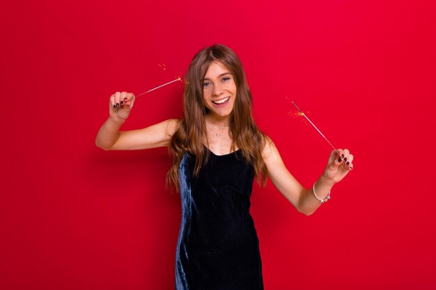 Brightful party event of joyful young happy woman in elegant black dress celebrating and having fun on isolated red background with sparklers.