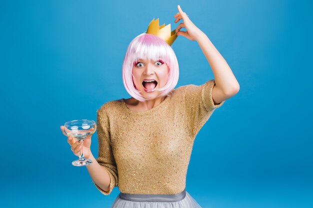 Brightful party time of funny young woman with champagne, golden crown on head having fun . Cut pink hair, expressing happiness, astonished, new year party, carnival.