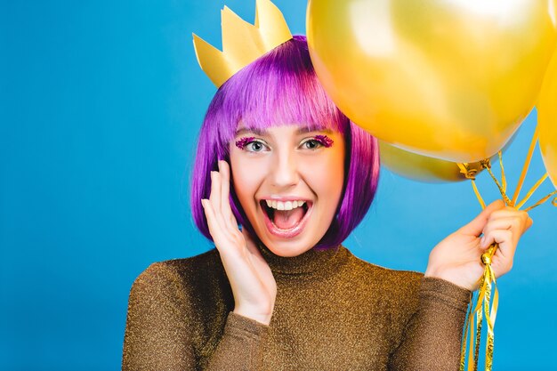 Brightful positive emotions at celebrating new year, birthday party of funny joyful young woman with cut purple hair . Golden balloons, crown on head, luxury dress, happiness.