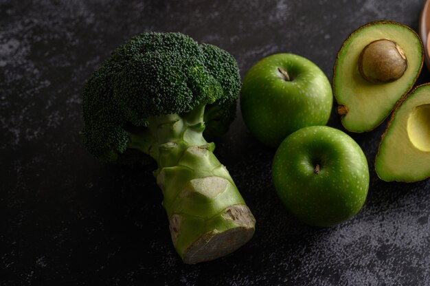 Broccoli, Apple, and avocado on a black cement floor.