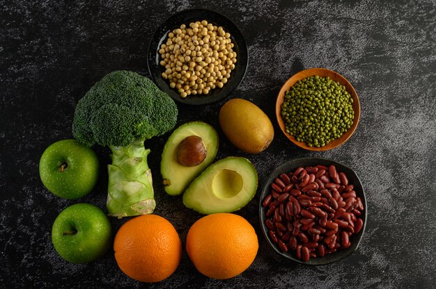 Broccoli, Apple, orange, kiwi, Legumes, and avocado on a black cement floor.