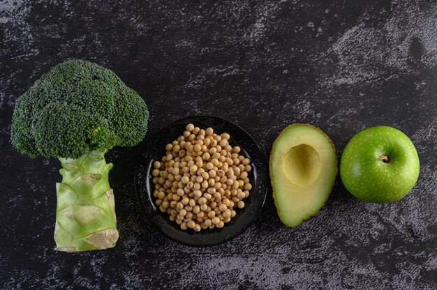 Broccoli, soy beans, Apple, and avocado on a black cement floor.