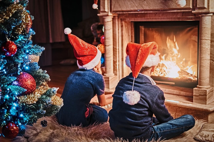 Free Photo brother and sister wearing santa's hats warming next to a fireplace in a living room decorated for christmas.