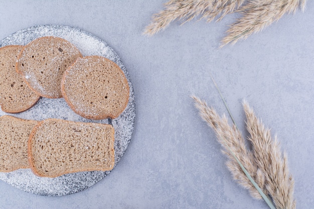 Brown bread slices on a flour covered board next to feather grass stalks on marble surface