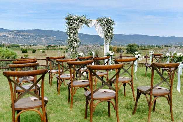 茶色のキアヴァリ椅子と晴れた日に白い花と緑の装飾が施された結婚式のアーチ