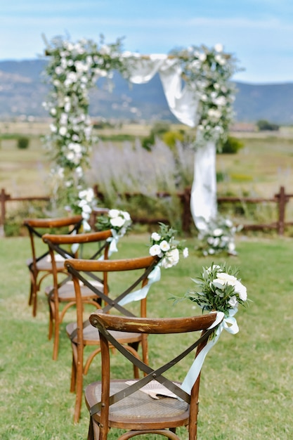 Brown chiavari chairs decorated with white eustomas bouquets on the grass and the decorated wedding archway on the background on the sunny day