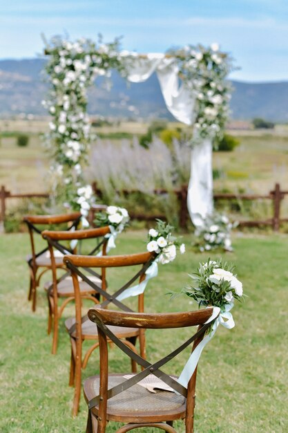 Стулья из коричневого чиавари, украшенные белыми букетами эустомы на траве и украшенной свадебной аркой на заднем плане в солнечный день