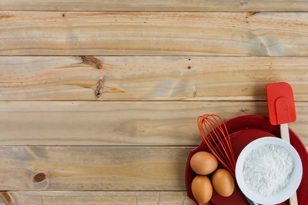 Brown eggs; flour and utensils on plate over wooden backdrop