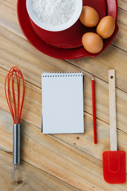 Brown eggs with flour on plate near spiral notepad; whisk; pencil and spatula over wooden surface