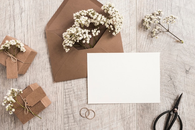 Brown envelope with baby's-breath flowers; gift boxes; wedding rings; scissor and white card on wooden background
