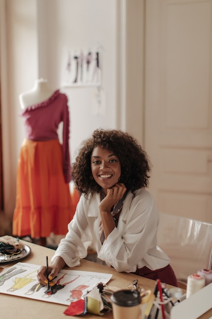 brown-eyed beautiful curly dark-skinned woman in stylish white blouse sits at desk, designs stylish clothes and smiles
