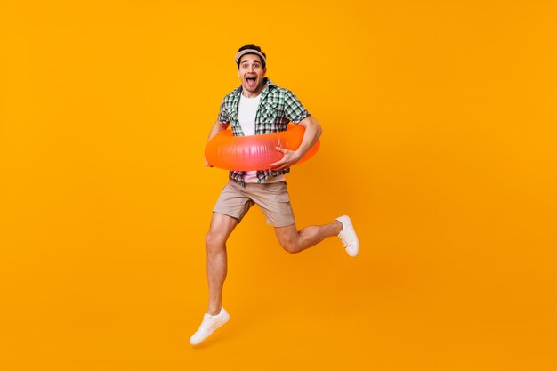 Кареглазый брюнет в бежевых шортах и зеленой футболке прыгает с надувным кругом на оранжевом пространстве.