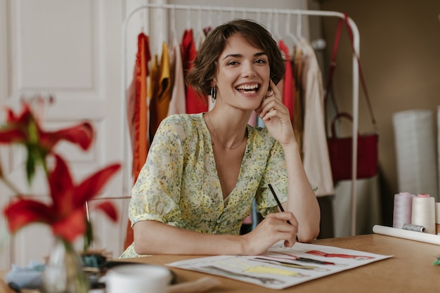 Brown-eyed curly brunette short-haired woman in floral trendy dress smiles, looks at camera, hoods pencil and designs new clothes