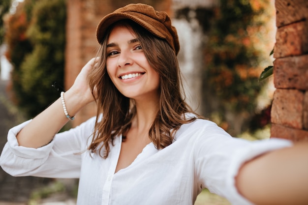 Ragazza dagli occhi marroni in berretto di velluto e camicetta bianca fa selfie sullo spazio del muro di mattoni e degli alberi.