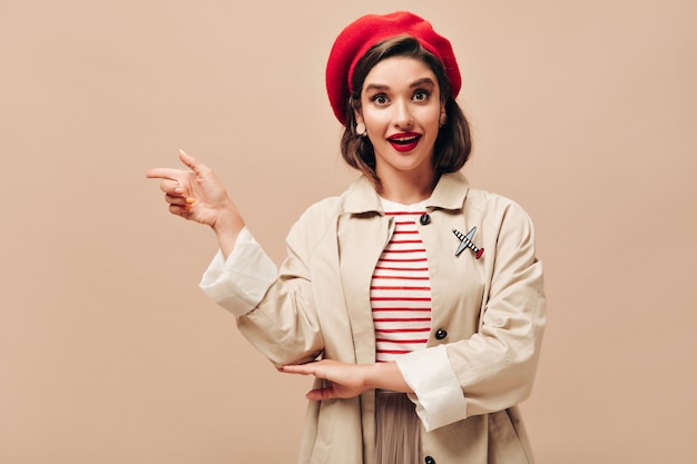 Free photo brown eyed lady in red beret and beige trench pointing to place for text. bright young woman in striped clothes posing for camera.