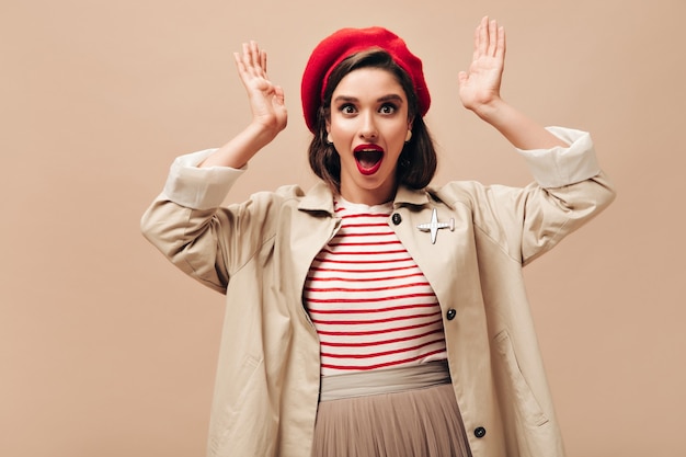 Brown eyed lady in trendy trench Surprised looks into camera. Shocked woman in striped sweater, beige skirt and coat posing on isolated background.