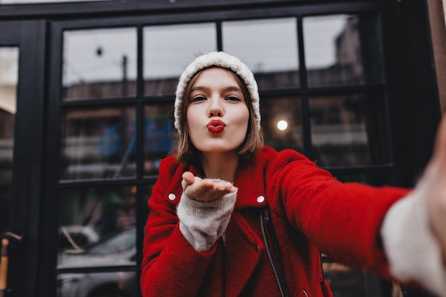 Brown-eyed lady in white hat blows kiss. Girl with red lipstick makes selfie against window.