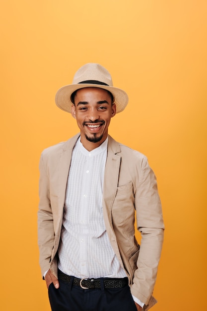 Brown-eyed man in shirt, beige jacket and hat smiling on orange wall