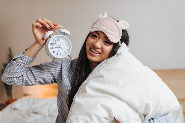 Foto gratuita donna dagli occhi marroni in pigiama blu e mascherina rosa per dormire posa con la sveglia sul letto