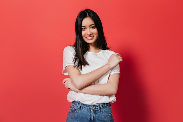 Brown-eyed woman in cotton top posing with smile on red wall