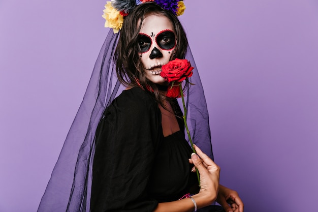 Brown-eyed woman dressed as widow demonstrates large red rose. Snapshot of brunette with unusual make-up in black outfit.