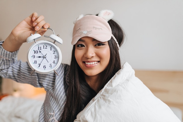 Brown-eyed woman in high spirits keeps alarm clock, smiles and looks into front