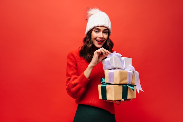 Brown-eyed woman in red sweater and white hat opens gift boxes