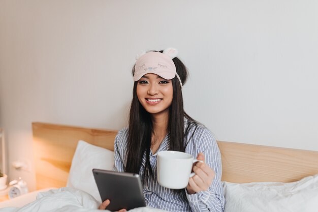 タブレットとカップでベッドに横たわっている間、茶色の目の女性は微笑んで正面を見る