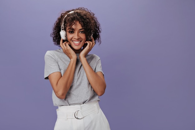 Brown-eyed woman in white headphones smiling over purple wall
