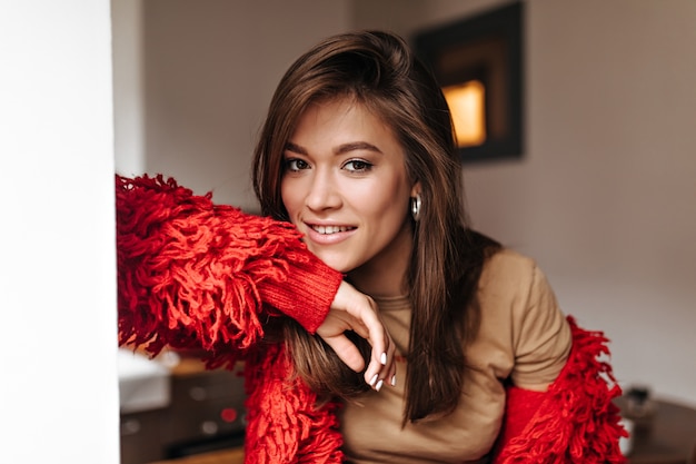 Brown-eyed woman with natural makeup looking at camera. Stylish lady in red jacket and light t-shirt looks into camera against background of room.