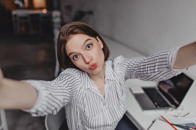 Brown-eyed young business woman makes selfie with funny facial expression against white table and laptop.