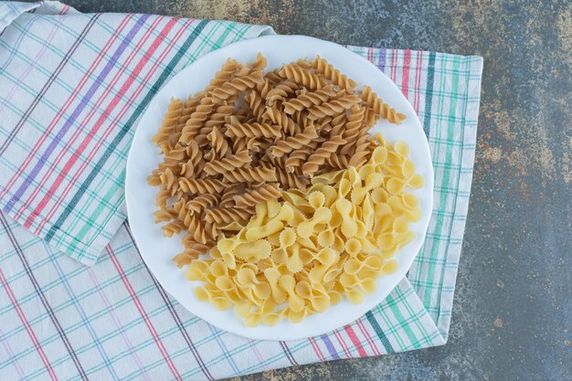 Brown fusilli pasta and farfalle pastas in the bowl on the towel, on the marble background.