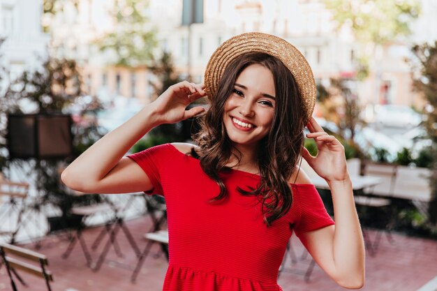 밀짚 모자에 포즈 갈색 머리 여성 모델입니다. 도시에 서있는 빨간 드레스에 좋은 기분 좋은 여자의 야외 사진