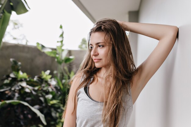 Brown-haired girl posing with dreamy face expression near white wall. Pensive caucasian lady touching her long hair and looking away.