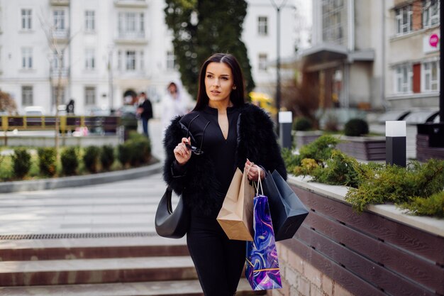 A brown-haired woman wearing black wear,  holds colourful, patterned shopping bags during a successful shopping spree. Walking outside, she is enjoying the warmth of a day